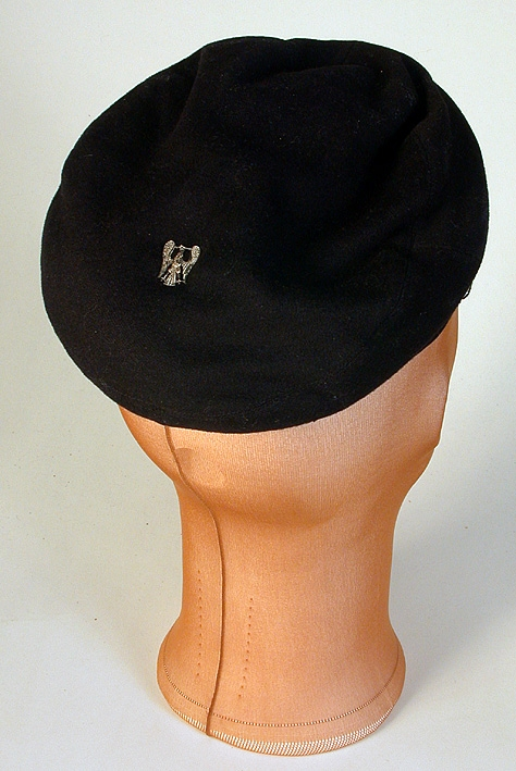 Sydd i svart ylle med lackerad skärm. Mösskanten med svart sammetsband med broderade blad mittfram.Vit snodd runt mössan. På kullen fastsydd en värmlandsörn av vitmetall. Svettsrem av brunt konstläder, fodrat med kyprat bomullstyg i gult och blått. På svettsremmen stansade små hål. P G R