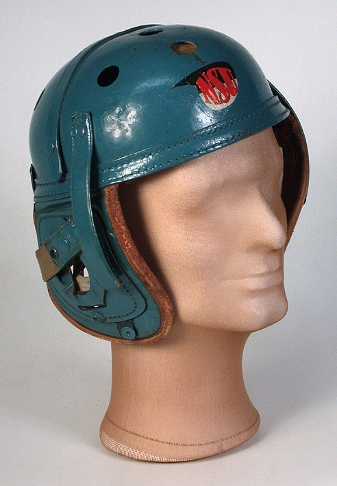 Ytterhölje bestående av en halvklotsformad kupa av turkosfärgat, hårt konstmaterial på insidan klädd med läder som på baksidan är stoppad med filt. På två motsatta sidor av kupans nederkant sitter två stycken, även de på utsidan klädda med det hårda konstmaterialet, på insidan med det stoppade lädret.  När kupan sätts på huvudet blir dessa som skydd för öron och kinder. På kupans baksida ett liknande stycke som skydd för nacke. På kupans ovansida, tio stycken ventilationshål vardera med diameter 20 mm. På framsidan står bokstäverna NSU i rött på en svart-vit bakgrund. Med ett ca 370 mm långt resårband med knappar i vardera ände som fästs på hjälmen, hålls hjälmen fast på huvudet. På insidan står stämplat: "Wilson Ath. Goods (oläsligt) Co. Chicago, Ill. 7 1/4.
