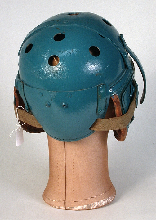 Ytterhölje bestående av en halvklotsformad kupa av turkosfärgat, hårt konstmaterial på insidan klädd med läder som på baksidan är stoppad med filt. På två motsatta sidor av kupans nederkant sitter två stycken, även de på utsidan klädda med det hårda konstmaterialet, på insidan med det stoppade lädret.  När kupan sätts på huvudet blir dessa som skydd för öron och kinder. På kupans baksida ett liknande stycke som skydd för nacke. På kupans ovansida, tio stycken ventilationshål vardera med diameter 20 mm. På framsidan står bokstäverna NSU i rött på en svart-vit bakgrund. Med ett ca 370 mm långt resårband med knappar i vardera ände som fästs på hjälmen, hålls hjälmen fast på huvudet. På insidan står stämplat: "Wilson Ath. Goods (oläsligt) Co. Chicago, Ill. 7 1/4.