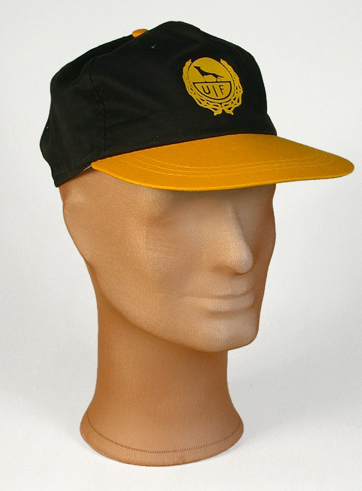 Keps i svart tyg. Skärm i hård papp klädd med klargult tyg på ovansidan och limegrönt tyg på undersidan. Klargul klädd knapp på kepsens topp. Mitt fram en cirkel med gult lagerbladsliknande mönster i hästskoform,  tryck med text  UIF samt hund eller varg siluett. Kepsen fodrad med vitt tyg mitt fram, vitt svettband. Mitt bak byglar för storleksändring i plast,