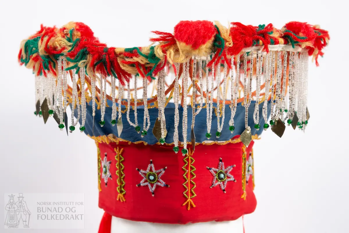 Krone til jonsokbryllup. Perlekrone med trøsk av papp. 
Utsida er dekka med et rødt syntetisk stoff. Innsida er dekka med ubleka bomullslerret, og er polstret med bomullsvatt. Det røde stoffet er perlebrodert, vekselsvis med stjerner og striper. Øverst midt bak er det sydd på to brede, røde bånd av samme stoff som det er festa rundt trøsken.
En liten spiss med mønstrete ullstoff med innvevd blomstermønster er sydd på i enden på hver av båndene, disse er kanta med perler.
Knytebånd av grovt, rødt bomullsbånd. Over trøsken sitter en smal bord med gule perler, deretter er festa et blått syntetisk bånd.
Begge deler støtta opp av spiler, som er dekka med et syntetisk oransje stoff. Øverst er spilene dekka med et hvitt skråbånd. Til øverste kant er festa dusker av ullgarn i rødt, gult og grønt.
Til øverste kant er det festa noen perlefrynser, noen er dekorert med en større rombeforma metallbit i enden, mens andre har en større grønn perle.
Eier har fått opplyst at dette skal stamme fra en gammel lampeskjerm. Krona har også en rad gule perler rett over det blå båndet.
All synlig søm er gjort for hånd.
Produsent laget utstyret sammen med sine barn ca. 1910-1920. Trolig brukt til 1970-åra.