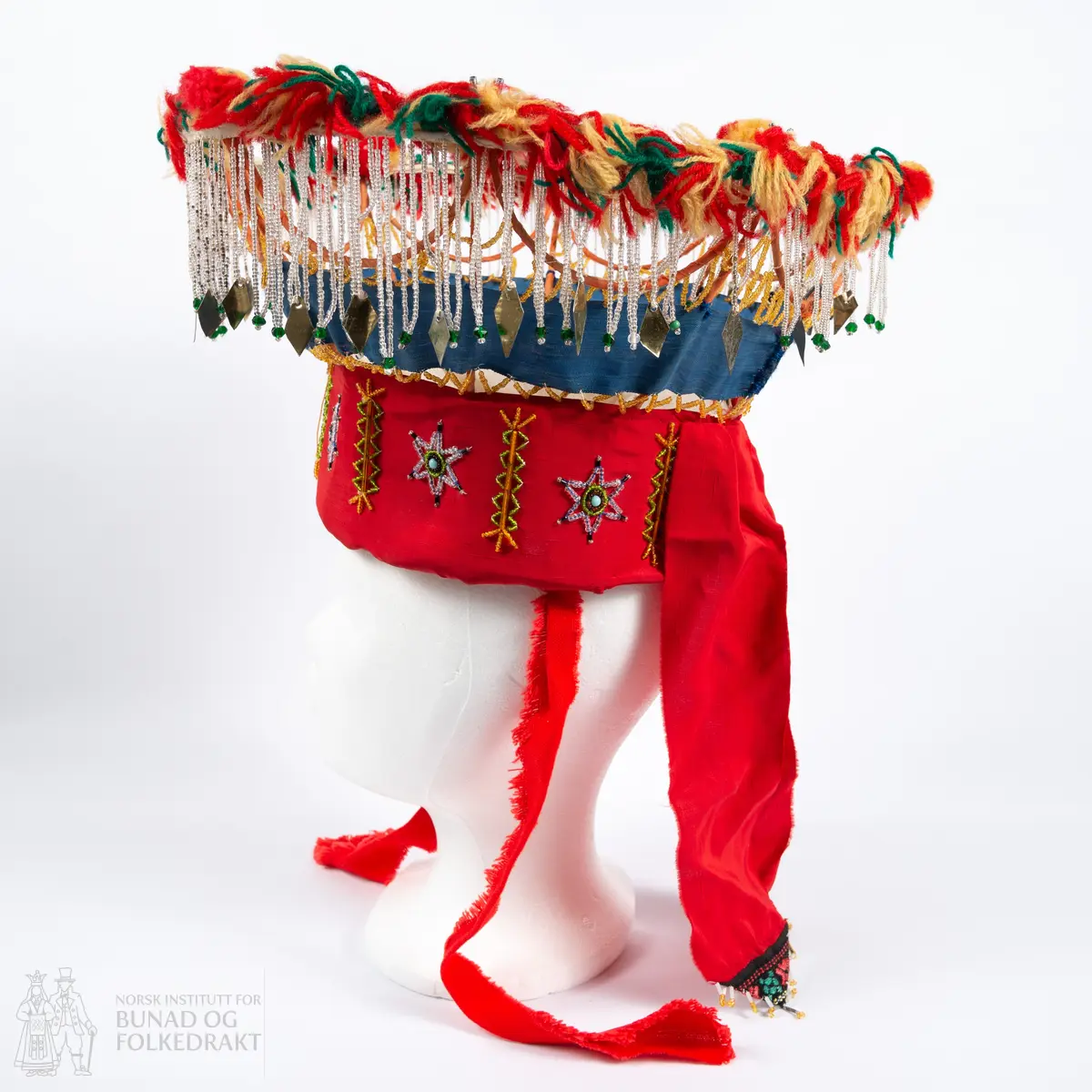 Krone til jonsokbryllup. Perlekrone med trøsk av papp. 
Utsida er dekka med et rødt syntetisk stoff. Innsida er dekka med ubleka bomullslerret, og er polstret med bomullsvatt. Det røde stoffet er perlebrodert, vekselsvis med stjerner og striper. Øverst midt bak er det sydd på to brede, røde bånd av samme stoff som det er festa rundt trøsken.
En liten spiss med mønstrete ullstoff med innvevd blomstermønster er sydd på i enden på hver av båndene, disse er kanta med perler.
Knytebånd av grovt, rødt bomullsbånd. Over trøsken sitter en smal bord med gule perler, deretter er festa et blått syntetisk bånd.
Begge deler støtta opp av spiler, som er dekka med et syntetisk oransje stoff. Øverst er spilene dekka med et hvitt skråbånd. Til øverste kant er festa dusker av ullgarn i rødt, gult og grønt.
Til øverste kant er det festa noen perlefrynser, noen er dekorert med en større rombeforma metallbit i enden, mens andre har en større grønn perle.
Eier har fått opplyst at dette skal stamme fra en gammel lampeskjerm. Krona har også en rad gule perler rett over det blå båndet.
All synlig søm er gjort for hånd.
Produsent laget utstyret sammen med sine barn ca. 1910-1920. Trolig brukt til 1970-åra.