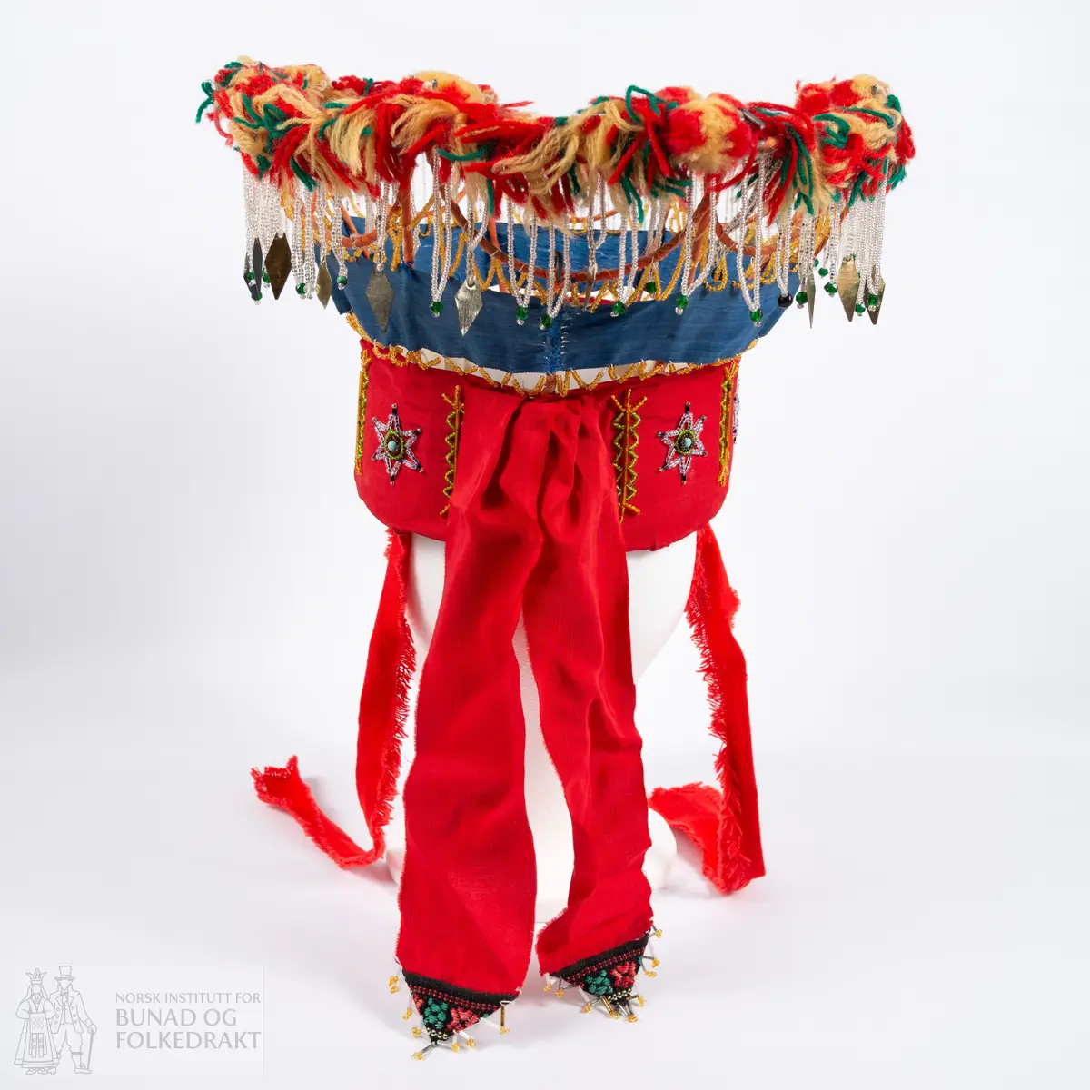 Krone til jonsokbryllup. Perlekrone med trøsk av papp. 
Utsida er dekka med et rødt syntetisk stoff. Innsida er dekka med ubleka bomullslerret, og er polstret med bomullsvatt. Det røde stoffet er perlebrodert, vekselsvis med stjerner og striper. Øverst midt bak er det sydd på to brede, røde bånd av samme stoff som det er festa rundt trøsken.
En liten spiss med mønstrete ullstoff med innvevd blomstermønster er sydd på i enden på hver av båndene, disse er kanta med perler.
Knytebånd av grovt, rødt bomullsbånd. Over trøsken sitter en smal bord med gule perler, deretter er festa et blått syntetisk bånd.
Begge deler støtta opp av spiler, som er dekka med et syntetisk oransje stoff. Øverst er spilene dekka med et hvitt skråbånd. Til øverste kant er festa dusker av ullgarn i rødt, gult og grønt.
Til øverste kant er det festa noen perlefrynser, noen er dekorert med en større rombeforma metallbit i enden, mens andre har en større grønn perle.
Eier har fått opplyst at dette skal stamme fra en gammel lampeskjerm. Krona har også en rad gule perler rett over det blå båndet.
All synlig søm er gjort for hånd.
Produsent laget utstyret sammen med sine barn ca. 1910-1920. Trolig brukt til 1970-åra.