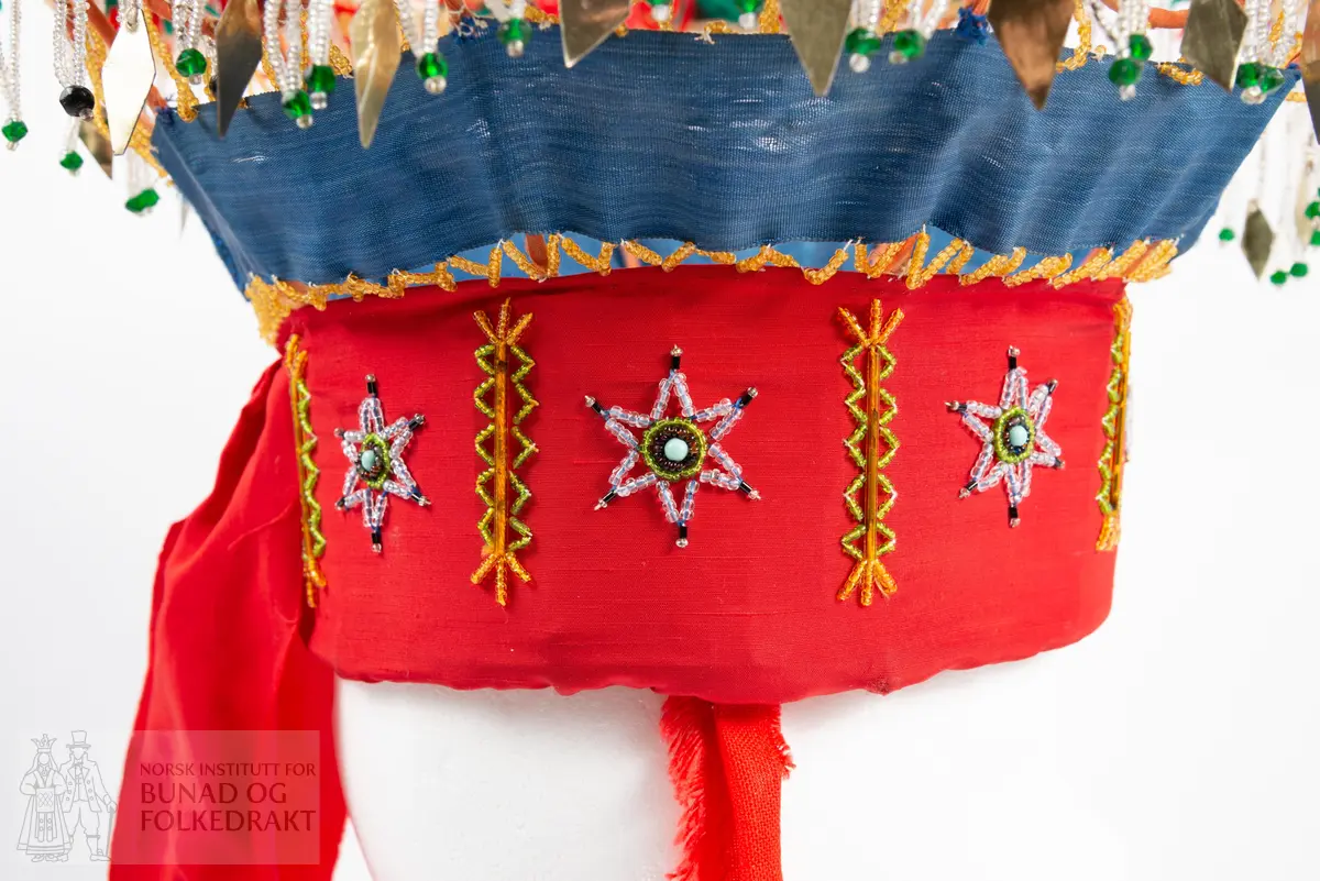 Krone til jonsokbryllup. Perlekrone med trøsk av papp. 
Utsida er dekka med et rødt syntetisk stoff. Innsida er dekka med ubleka bomullslerret, og er polstret med bomullsvatt. Det røde stoffet er perlebrodert, vekselsvis med stjerner og striper. Øverst midt bak er det sydd på to brede, røde bånd av samme stoff som det er festa rundt trøsken.
En liten spiss med mønstrete ullstoff med innvevd blomstermønster er sydd på i enden på hver av båndene, disse er kanta med perler.
Knytebånd av grovt, rødt bomullsbånd. Over trøsken sitter en smal bord med gule perler, deretter er festa et blått syntetisk bånd.
Begge deler støtta opp av spiler, som er dekka med et syntetisk oransje stoff. Øverst er spilene dekka med et hvitt skråbånd. Til øverste kant er festa dusker av ullgarn i rødt, gult og grønt.
Til øverste kant er det festa noen perlefrynser, noen er dekorert med en større rombeforma metallbit i enden, mens andre har en større grønn perle.
Eier har fått opplyst at dette skal stamme fra en gammel lampeskjerm. Krona har også en rad gule perler rett over det blå båndet.
All synlig søm er gjort for hånd.
Produsent laget utstyret sammen med sine barn ca. 1910-1920. Trolig brukt til 1970-åra.