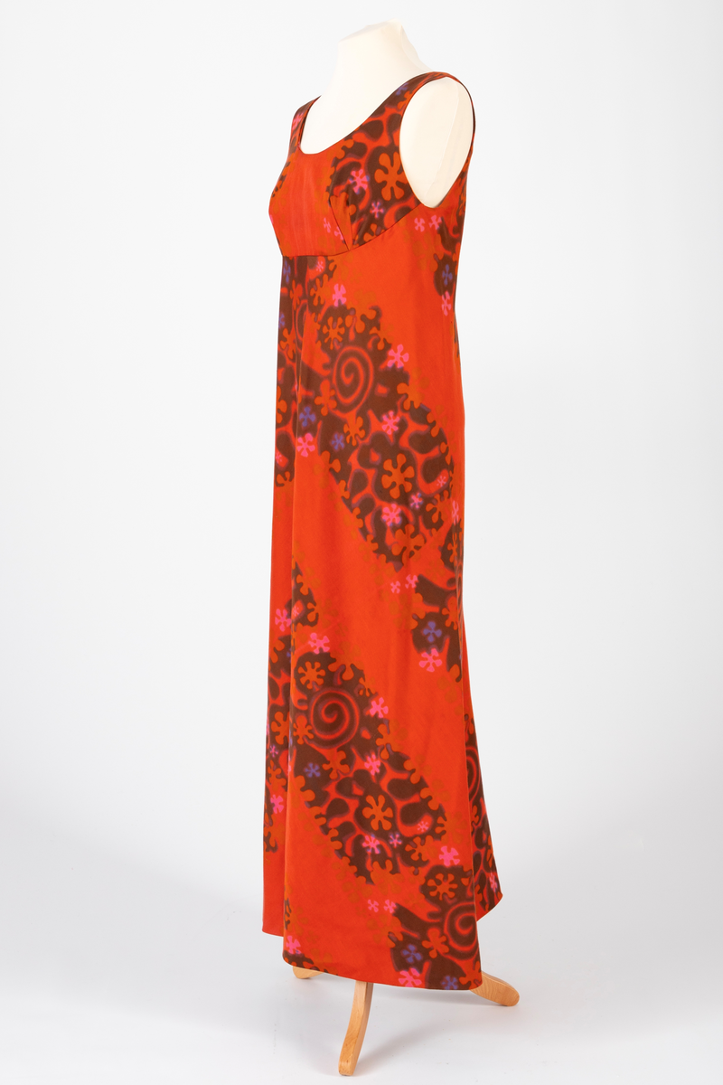 Lang, ermeløs kjole i rødoransje mønstertrykket, satengvevd bomullsstoff. Kjolen er lett innsvinget med høyt liv og utskrådd skjørt. Lav, rundet utringning foran og v-formet utringning bak. Kjolen har oransje silkefôr og glidelås i ryggen.