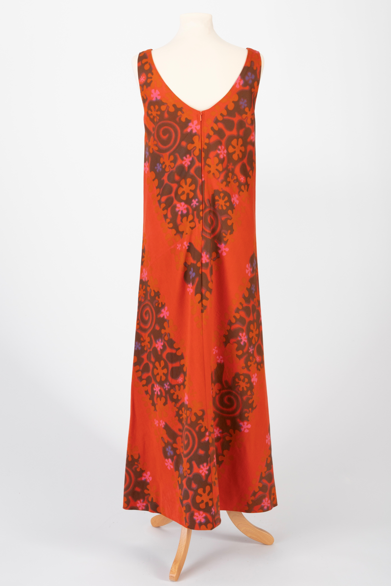 Lang, ermeløs kjole i rødoransje mønstertrykket, satengvevd bomullsstoff. Kjolen er lett innsvinget med høyt liv og utskrådd skjørt. Lav, rundet utringning foran og v-formet utringning bak. Kjolen har oransje silkefôr og glidelås i ryggen.