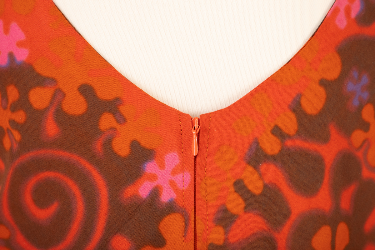 Lang, ermeløs kjole i rødoransje mønstertrykket, satengvevd bomullsstoff. Kjolen er lett innsvinget med høyt liv og utskrådd skjørt. Lav, rundet utringning foran og v-formet utringning bak. Kjolen har oransje silkefôr og glidelås i ryggen.