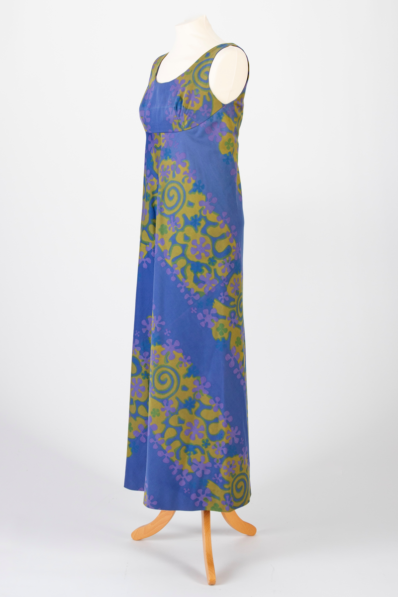 Lang, ermeløs kjole i blålilla mønstertrykket, satengvevd bomullsstoff. Kjolen er lett innsvinget med høyt liv og utskrådd skjørt. Lav, rundet utringning foran og v-formet utringning bak. Kjolen har mørkeblått silkefôr og glidelås i ryggen.