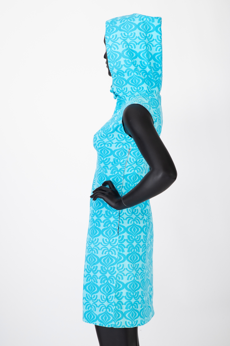 Knekort, ermeløs sommerkjole med hette. Turkis bomull med tettstilt, stilisert mønstertrykk. Mønsteret framstår som nonfigurativ, men minner om en stor løk med bladformasjon. Kjolen har A-linje form, glidelås i venstre side og stofftrukne knapper i halssplitt. Helfôret.