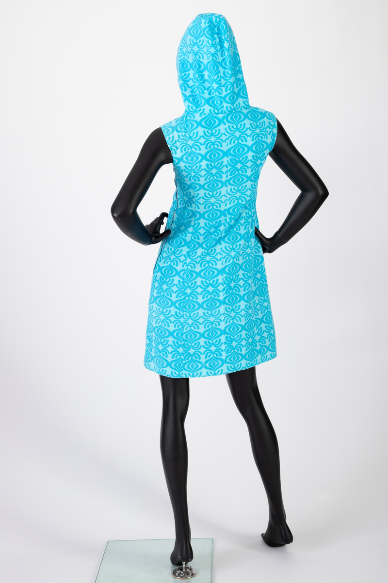 Knekort, ermeløs sommerkjole med hette. Turkis bomull med tettstilt, stilisert mønstertrykk. Mønsteret framstår som nonfigurativ, men minner om en stor løk med bladformasjon. Kjolen har A-linje form, glidelås i venstre side og stofftrukne knapper i halssplitt. Helfôret.