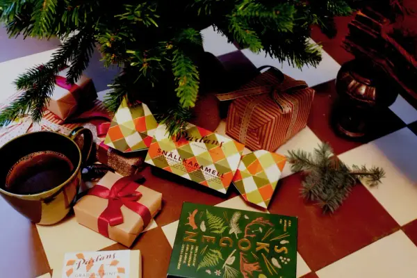 Paket i olika former och färger som ligger under en gran på ett rödvitrutigt golv.