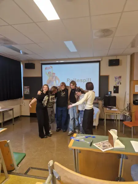 Flere personer holder hverandre rundt skuldrene foran i et klasserom. På skjermen bak dem ses ordet Rollespill.