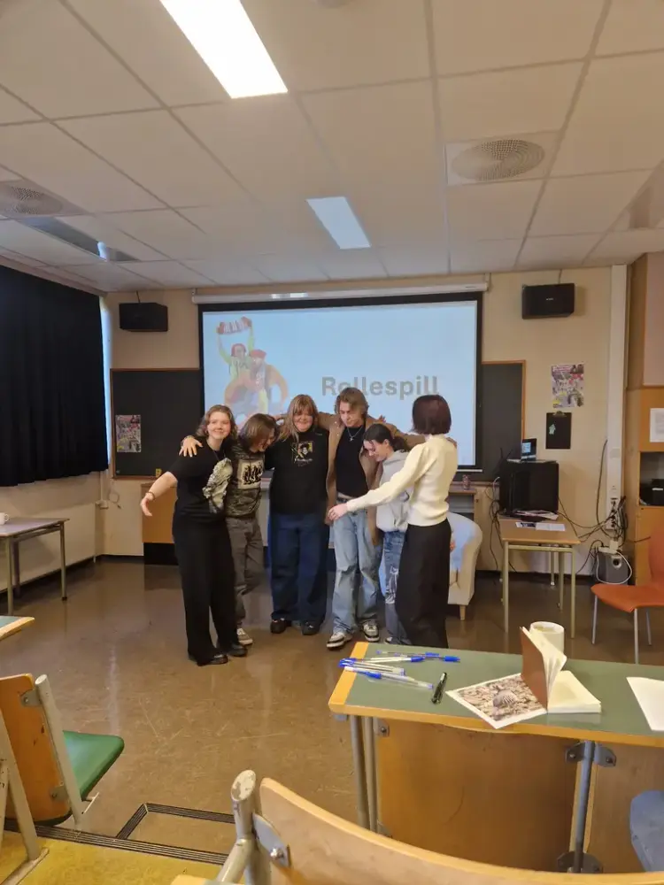 Flere personer holder hverandre rundt skuldrene foran i et klasserom. På skjermen bak dem ses ordet Rollespill.