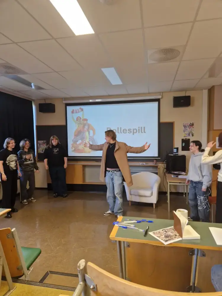 En person i beige frakk står med åpne armer foran tavla i et klasserom. Flere elever står rundt og ser på.