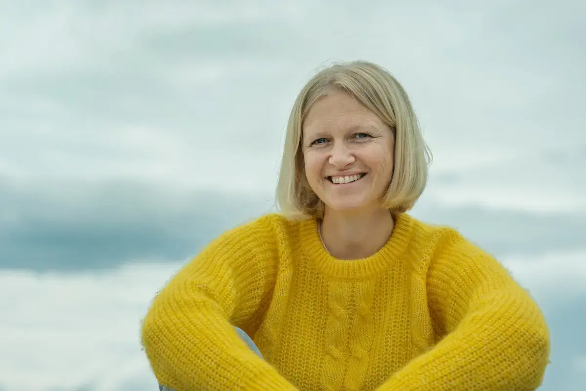Maria Parr i gul strikkegenser sitjande med ein tilskya himmel som bakgrunn