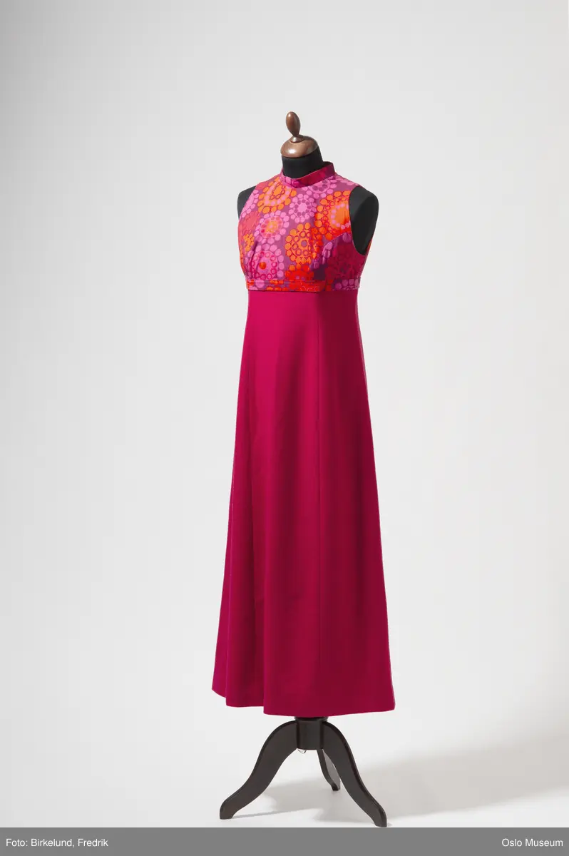 Lang ermeløs kjole i rød ull. Foret. Blomstret bærestykke i bomull. Glidelås i ryggen og hempe bak.