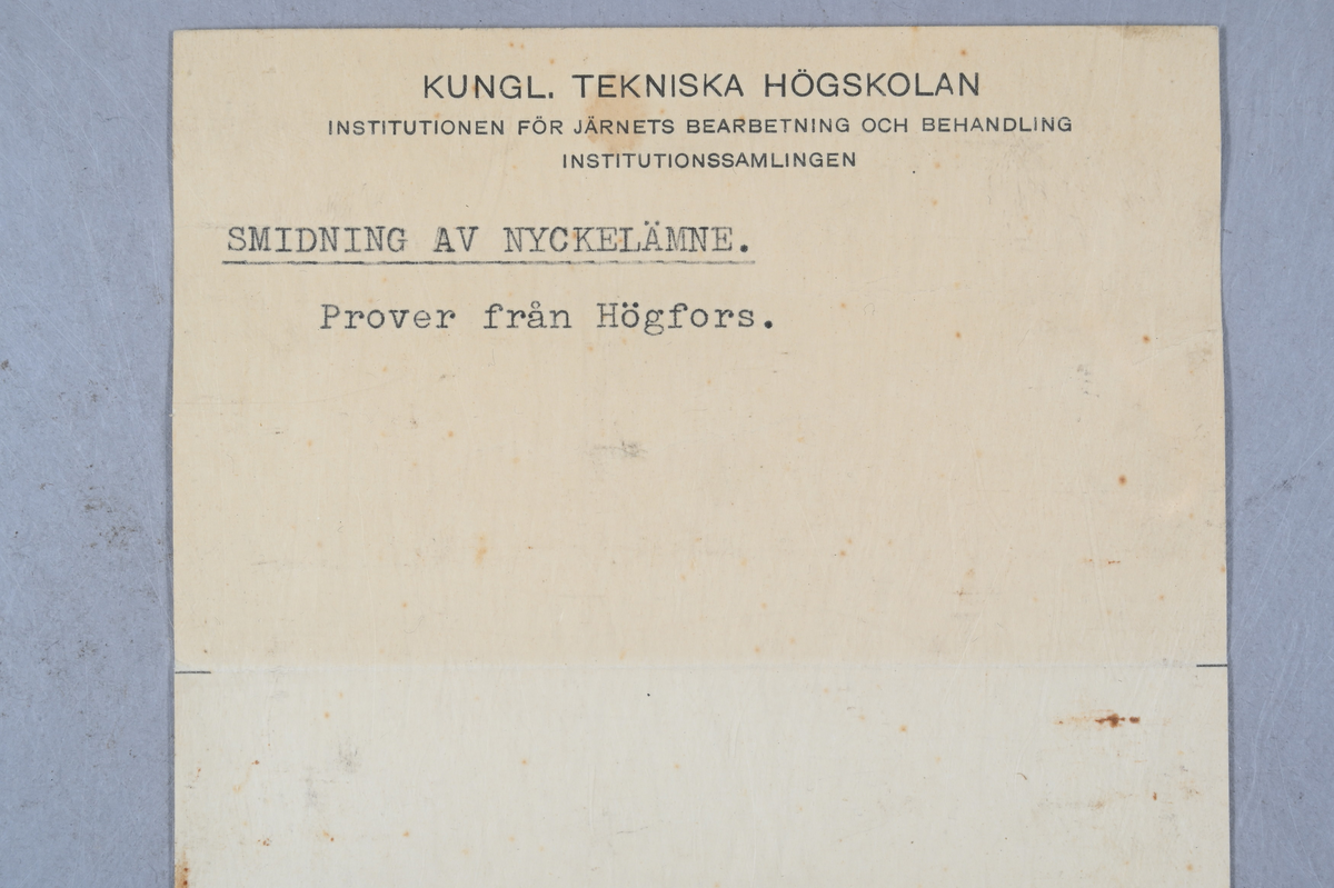 Två prover på nyckelämnen av järn. Märkning på ena provet med stämpel: "Högfors". Ett av proverna visar smidning av nyckelämne. Båda proverna från Högfors. Tillhörande lapp med samma notering.