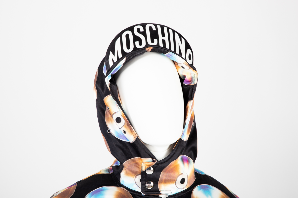 Vid jakke av ganske blankt, svart materiale, med påtrykt mønster i form av tettstilte, metallskimrende disker/CD'er/DVD'er. Jakken har blusongfasong, dvs. holdt inn i livhøyde med påsatt elastikk i nederkant. Tilsvarende elastikk i mansjetter på de lange ermene. Lukkes midt foran med kraftig glidelås over stukket og vattert belegg. Skråstilte lommer med belegg og klaff på hvert forstykke. Klaffene lukket med blank trykknapp. I tillegg påsydd lomme med glidelås på yttersiden av venstre erme og med utvendig rom for to blyanter. Jakken har også to innvendige lommer, én på hvert forstykke. De innvendige lommene er lukket med trykknapp. Måneformet kile isydd under hvert erme. Disse er utstyrt med tre luftehull av metallmaljer i ytterstoff. Det samme gjelder tre maljer i hetten ved hvert øre. Jakken er helfôret med svart materiale, ivevd merkenavnet Moschino, tett-i-tett. Hetten er utstyrt med stiv "negl", dvs. "skygge"/brem, som er nestet opp, slik at man kan lese navnet MOSCHINO trykket i hvitt mot svart bakgrunn. Baksiden/oversiden av "skyggen" er trukket med det ytre stoffet. Hetten lukkes med en forlengelse midt foran, som fungerer som en overlappende halsklaff og lukkes med to vertikalstilte trykknapper.