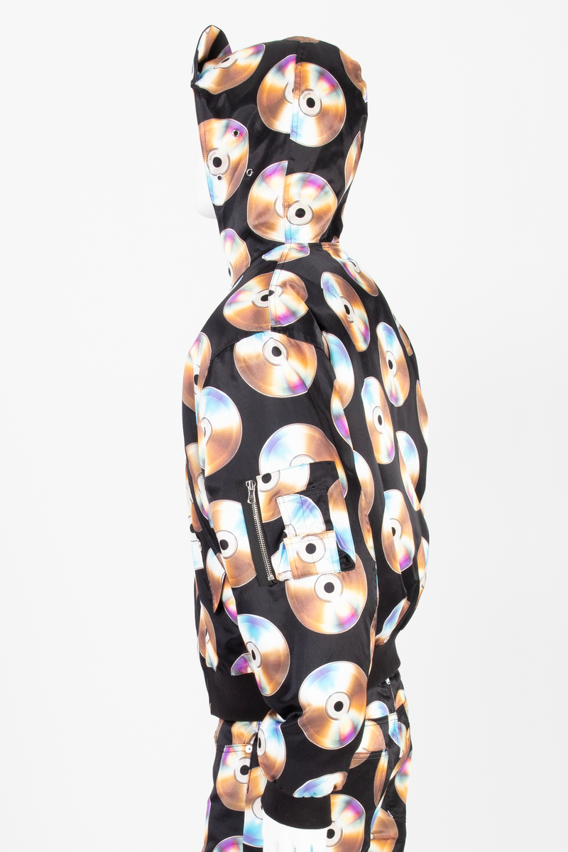 Vid jakke av ganske blankt, svart materiale, med påtrykt mønster i form av tettstilte, metallskimrende disker/CD'er/DVD'er. Jakken har blusongfasong, dvs. holdt inn i livhøyde med påsatt elastikk i nederkant. Tilsvarende elastikk i mansjetter på de lange ermene. Lukkes midt foran med kraftig glidelås over stukket og vattert belegg. Skråstilte lommer med belegg og klaff på hvert forstykke. Klaffene lukket med blank trykknapp. I tillegg påsydd lomme med glidelås på yttersiden av venstre erme og med utvendig rom for to blyanter. Jakken har også to innvendige lommer, én på hvert forstykke. De innvendige lommene er lukket med trykknapp. Måneformet kile isydd under hvert erme. Disse er utstyrt med tre luftehull av metallmaljer i ytterstoff. Det samme gjelder tre maljer i hetten ved hvert øre. Jakken er helfôret med svart materiale, ivevd merkenavnet Moschino, tett-i-tett. Hetten er utstyrt med stiv "negl", dvs. "skygge"/brem, som er nestet opp, slik at man kan lese navnet MOSCHINO trykket i hvitt mot svart bakgrunn. Baksiden/oversiden av "skyggen" er trukket med det ytre stoffet. Hetten lukkes med en forlengelse midt foran, som fungerer som en overlappende halsklaff og lukkes med to vertikalstilte trykknapper.