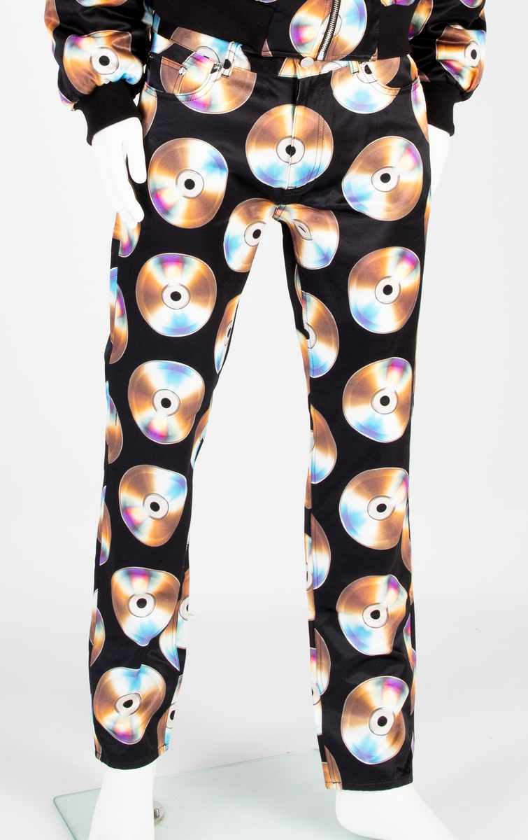 Lavtsittende, slank herrebukse av ganske blankt, svart materiale, med påtrykt mønster i form av tettstilte, metallskimrende disker/CD'er/DVD'er, noen av dem påtrykt navnet Moschino. Gylf lukket med glidelås, linning med beltestoler og lukket med knapp og knapphull. Jeanslomme på hvert forstykke, den høyre har i tillegg "pengelomme". Alle lommer på forsiden er forsterket med metallnagler og har bomullsfôr. Baksiden har bærestykke opp mot linningen og påsydde, åpne lommer på hvert bakstykke.