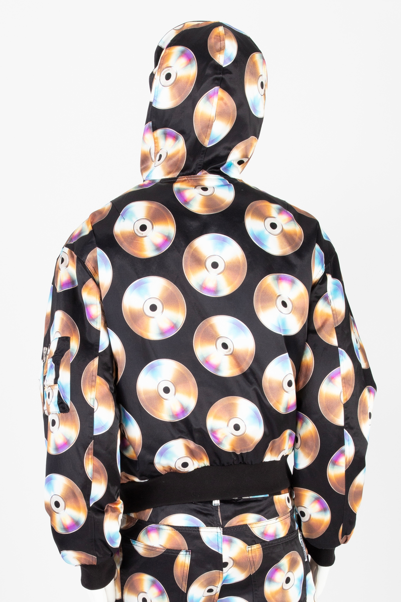 Vid jakke av ganske blankt, svart materiale, med påtrykt mønster i form av tettstilte, metallskimrende disker/CD'er/DVD'er. Jakken har blusongfasong, dvs. holdt inn i livhøyde med påsatt elastikk i nederkant. Tilsvarende elastikk i mansjetter på de lange ermene. Lukkes midt foran med kraftig glidelås over stukket og vattert belegg. Skråstilte lommer med belegg og klaff på hvert forstykke. Klaffene lukket med blank trykknapp. I tillegg påsydd lomme med glidelås på yttersiden av venstre erme og med utvendig rom for to blyanter. Jakken har også to innvendige lommer, én på hvert forstykke. De innvendige lommene er lukket med trykknapp. Måneformet kile isydd under hvert erme. Disse er utstyrt med tre luftehull av metallmaljer i ytterstoff. Det samme gjelder tre maljer i hetten ved hvert øre. Jakken er helfôret med svart materiale, ivevd merkenavnet Moschino, tett-i-tett. Hetten er utstyrt med stiv "negl", dvs. "skygge"/brem, som er nestet opp, slik at man kan lese navnet MOSCHINO trykket i hvitt mot svart bakgrunn. Baksiden/oversiden av "skyggen" er trukket med det ytre stoffet. Hetten lukkes med en forlengelse midt foran, som fungerer som en overlappende halsklaff og lukkes med to vertikalstilte trykknapper.