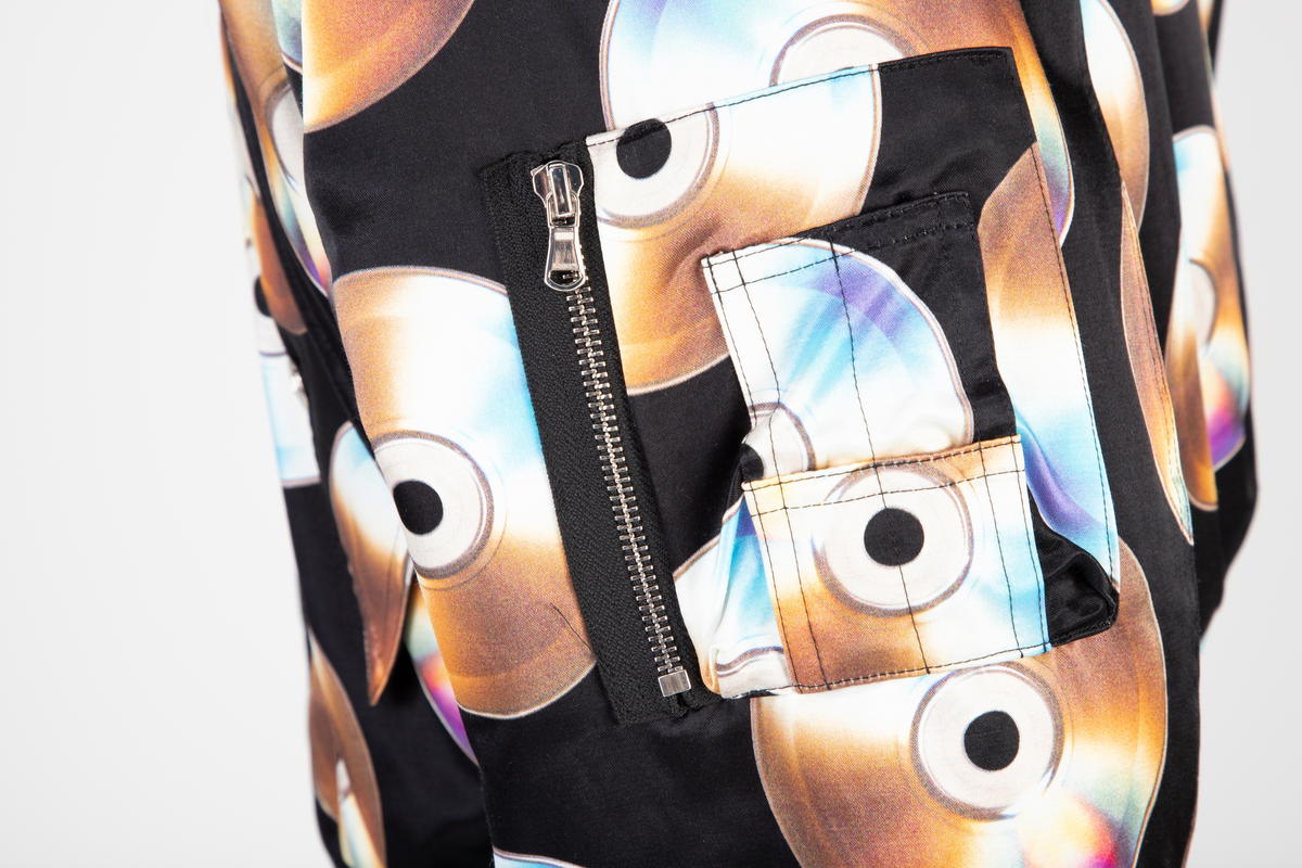 Vid jakke av ganske blankt, svart materiale, med påtrykt mønster i form av tettstilte, metallskimrende disker/CD'er/DVD'er. Jakken har blusongfasong, dvs. holdt inn i livhøyde med påsatt elastikk i nederkant. Tilsvarende elastikk i mansjetter på de lange ermene. Lukkes midt foran med kraftig glidelås over stukket og vattert belegg. Skråstilte lommer med belegg og klaff på hvert forstykke. Klaffene lukket med blank trykknapp. I tillegg påsydd lomme med glidelås på yttersiden av venstre erme og med utvendig rom for to blyanter. Jakken har også to innvendige lommer, én på hvert forstykke. De innvendige lommene er lukket med trykknapp. Måneformet kile isydd under hvert erme. Disse er utstyrt med tre luftehull av metallmaljer i ytterstoff. Det samme gjelder tre maljer i hetten ved hvert øre. Jakken er helfôret med svart materiale, ivevd merkenavnet Moschino, tett-i-tett. Hetten er utstyrt med stiv "negl", dvs. "skygge"/brem, som er nestet opp, slik at man kan lese navnet MOSCHINO trykket i hvitt mot svart bakgrunn. Baksiden/oversiden av "skyggen" er trukket med det ytre stoffet. Hetten lukkes med en forlengelse midt foran, som fungerer som en overlappende halsklaff og lukkes med to vertikalstilte trykknapper.