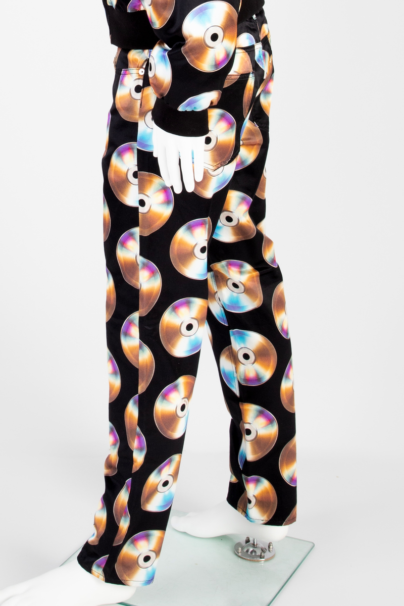 Lavtsittende, slank herrebukse av ganske blankt, svart materiale, med påtrykt mønster i form av tettstilte, metallskimrende disker/CD'er/DVD'er, noen av dem påtrykt navnet Moschino. Gylf lukket med glidelås, linning med beltestoler og lukket med knapp og knapphull. Jeanslomme på hvert forstykke, den høyre har i tillegg "pengelomme". Alle lommer på forsiden er forsterket med metallnagler og har bomullsfôr. Baksiden har bærestykke opp mot linningen og påsydde, åpne lommer på hvert bakstykke.