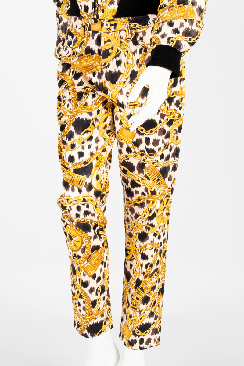 Lavtsittende, slank damebukse av ganske blankt materiale, med påtrykt mønster i form av tettstilte, gullfargede lenker, hengelåser og bærbare radioer/spillere, noen av dem påtrykt navnet Moschino, mot en bakgrunn med dyremønster. Gylf lukket med glidelås, linning med beltestoler og lukket med knapp og knapphull. Jeanslomme på hvert forstykke, den høyre har i tillegg "pengelomme". Alle lommer på forsiden er forsterket med metallnagler og har bomullsfôr. Baksiden har bærestykke opp mot linningen og påsydde, åpne lommer på hvert bakstykke.