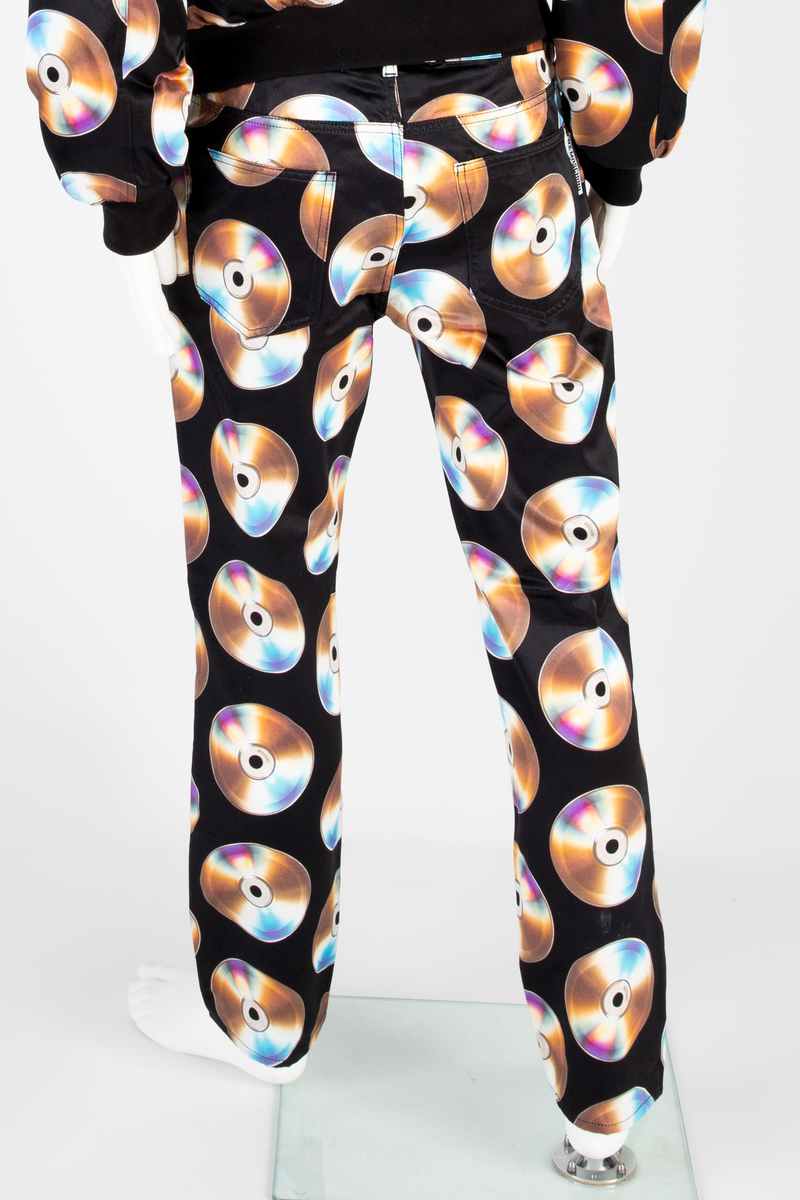 Lavtsittende, slank herrebukse av ganske blankt, svart materiale, med påtrykt mønster i form av tettstilte, metallskimrende disker/CD'er/DVD'er, noen av dem påtrykt navnet Moschino. Gylf lukket med glidelås, linning med beltestoler og lukket med knapp og knapphull. Jeanslomme på hvert forstykke, den høyre har i tillegg "pengelomme". Alle lommer på forsiden er forsterket med metallnagler og har bomullsfôr. Baksiden har bærestykke opp mot linningen og påsydde, åpne lommer på hvert bakstykke.