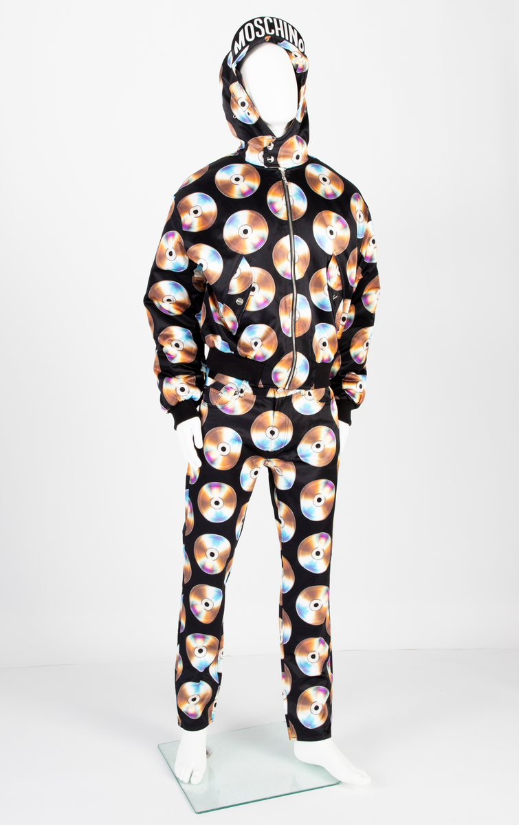 Lavtsittende, slank herrebukse av ganske blankt, svart materiale, med påtrykt mønster i form av tettstilte, metallskimrende disker/CD'er/DVD'er, noen av dem påtrykt navnet Moschino. Gylf lukket med glidelås, linning med beltestoler og lukket med knapp og knapphull. Jeanslomme på hvert forstykke, den høyre har i tillegg "pengelomme". Alle lommer på forsiden er forsterket med metallnagler og har bomullsfôr. Baksiden har bærestykke opp mot linningen og påsydde, åpne lommer på hvert bakstykke.