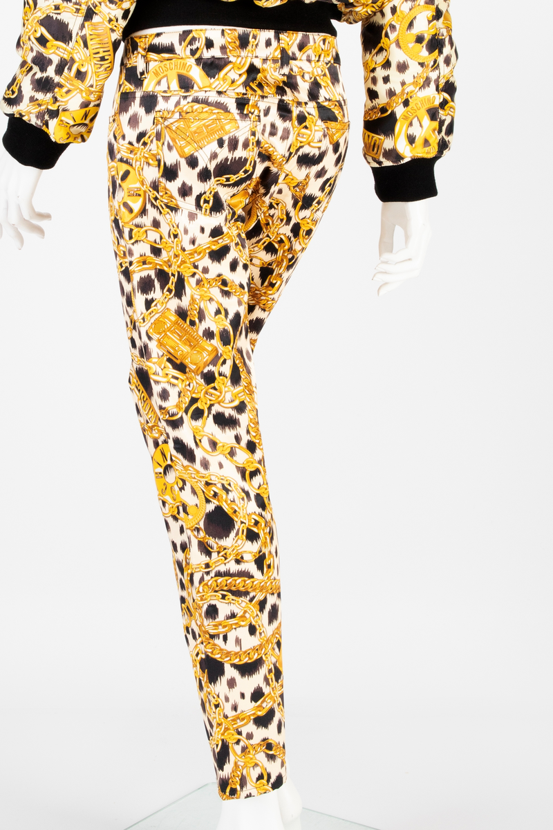 Lavtsittende, slank damebukse av ganske blankt materiale, med påtrykt mønster i form av tettstilte, gullfargede lenker, hengelåser og bærbare radioer/spillere, noen av dem påtrykt navnet Moschino, mot en bakgrunn med dyremønster. Gylf lukket med glidelås, linning med beltestoler og lukket med knapp og knapphull. Jeanslomme på hvert forstykke, den høyre har i tillegg "pengelomme". Alle lommer på forsiden er forsterket med metallnagler og har bomullsfôr. Baksiden har bærestykke opp mot linningen og påsydde, åpne lommer på hvert bakstykke.