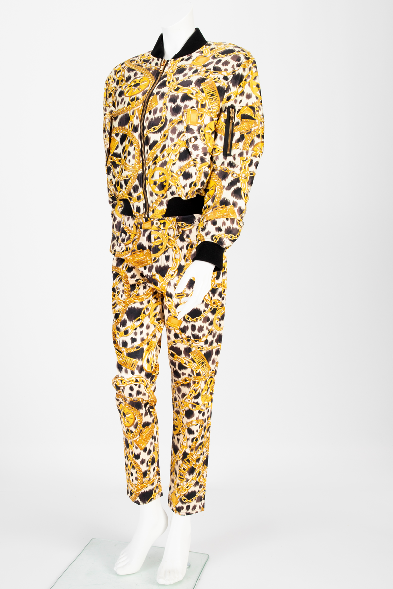 Lavtsittende, slank damebukse av ganske blankt materiale, med påtrykt mønster i form av tettstilte, gullfargede lenker, hengelåser og bærbare radioer/spillere, noen av dem påtrykt navnet Moschino, mot en bakgrunn med dyremønster. Gylf lukket med glidelås, linning med beltestoler og lukket med knapp og knapphull. Jeanslomme på hvert forstykke, den høyre har i tillegg "pengelomme". Alle lommer på forsiden er forsterket med metallnagler og har bomullsfôr. Baksiden har bærestykke opp mot linningen og påsydde, åpne lommer på hvert bakstykke.