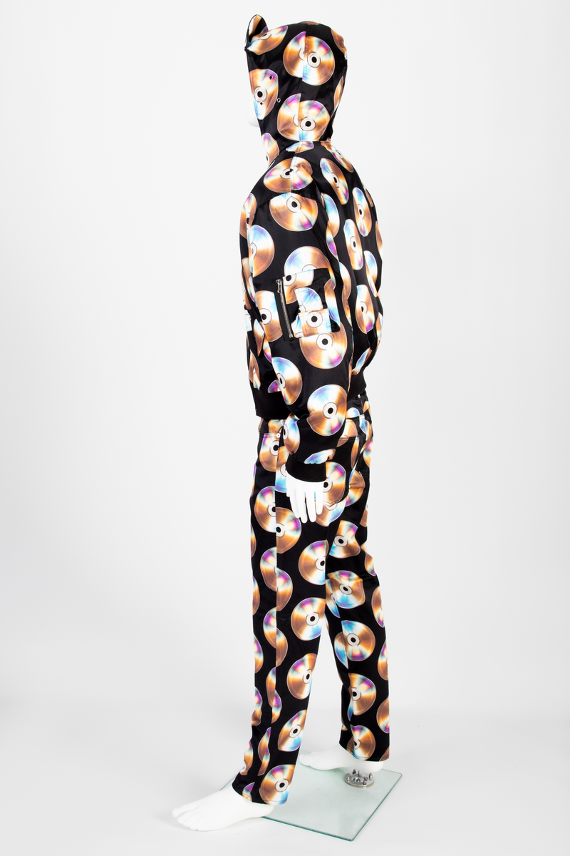 Lavtsittende, slank herrebukse av ganske blankt, svart materiale, med påtrykt mønster i form av tettstilte, metallskimrende disker/CD'er/DVD'er, noen av dem påtrykt navnet Moschino. Gylf lukket med glidelås, linning med beltestoler og lukket med knapp og knapphull. Jeanslomme på hvert forstykke, den høyre har i tillegg "pengelomme". Alle lommer på forsiden er forsterket med metallnagler og har bomullsfôr. Baksiden har bærestykke opp mot linningen og påsydde, åpne lommer på hvert bakstykke.