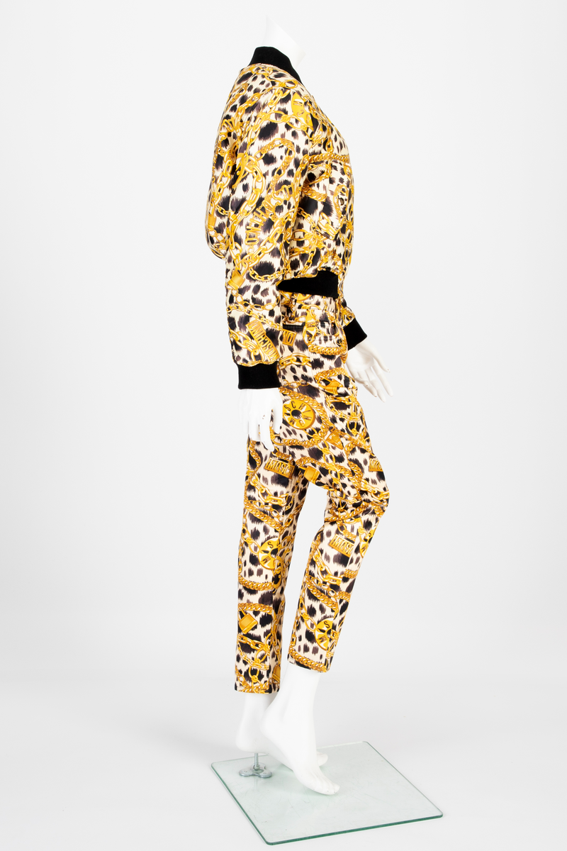 Lavtsittende, slank damebukse av ganske blankt materiale, med påtrykt mønster i form av tettstilte, gullfargede lenker, hengelåser og bærbare radioer/spillere, noen av dem påtrykt navnet Moschino, mot en bakgrunn med dyremønster. Gylf lukket med glidelås, linning med beltestoler og lukket med knapp og knapphull. Jeanslomme på hvert forstykke, den høyre har i tillegg "pengelomme". Alle lommer på forsiden er forsterket med metallnagler og har bomullsfôr. Baksiden har bærestykke opp mot linningen og påsydde, åpne lommer på hvert bakstykke.