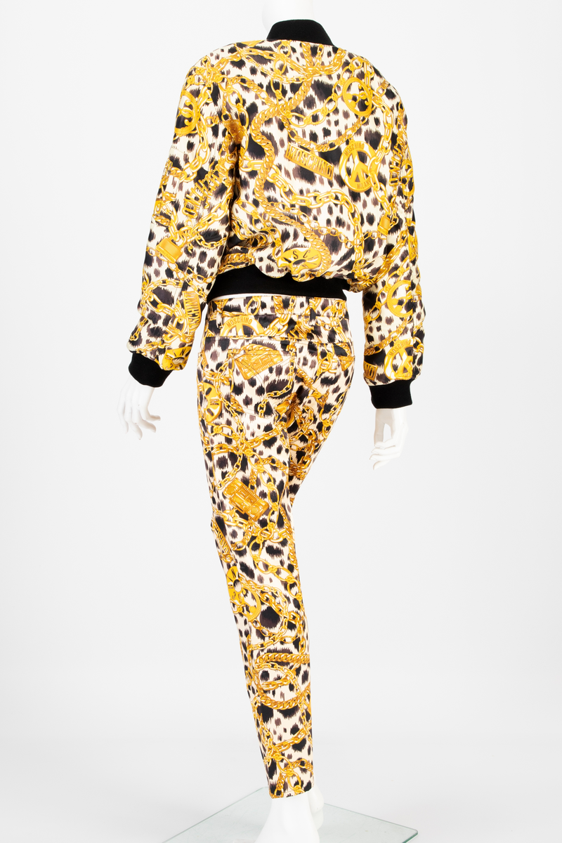 Lavtsittende, slank damebukse av ganske blankt materiale, med påtrykt mønster i form av tettstilte, gullfargede lenker, hengelåser og bærbare radioer/spillere, noen av dem påtrykt navnet Moschino, mot en bakgrunn med dyremønster. Gylf lukket med glidelås, linning med beltestoler og lukket med knapp og knapphull. Jeanslomme på hvert forstykke, den høyre har i tillegg "pengelomme". Alle lommer på forsiden er forsterket med metallnagler og har bomullsfôr. Baksiden har bærestykke opp mot linningen og påsydde, åpne lommer på hvert bakstykke.