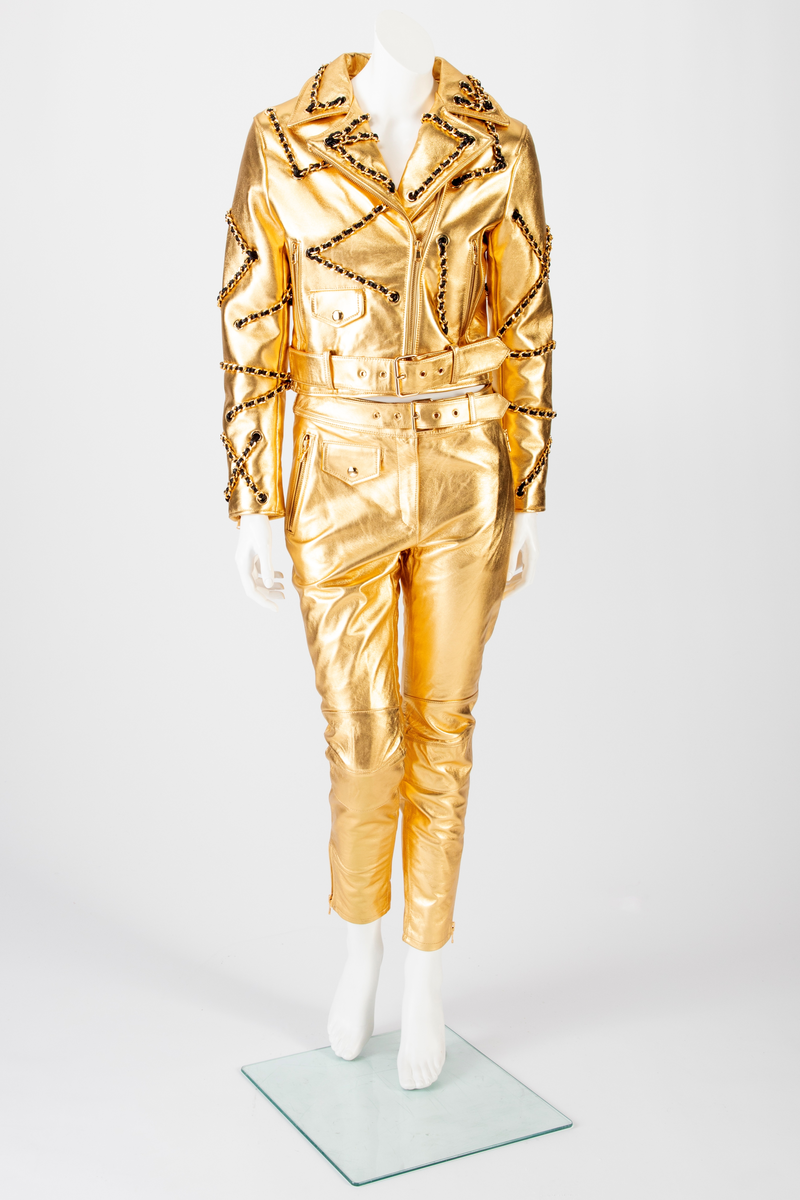 Skinnbukse, jeans-modell, med passepoilerte stikklommer med gullforgylt metallglidelås. På høyre side, nær linningen, en urlomme med klaff og gulltrykknapp. Gylf midt foran med gullforgylt glidelås og trykknapp i linningen. En passepoilert baklomme med gullforgylt glidelås på høyre side. På venstre side er lommen lik urlommen foran. Linningen har fem stikninger. Beltet kommer ut av linningen ved sidesømmen, Beltet har gullforgylt metallspenne og maljer. Formet over kneet. Gullforgylt glidelås med passepoiler ved fotvidden.