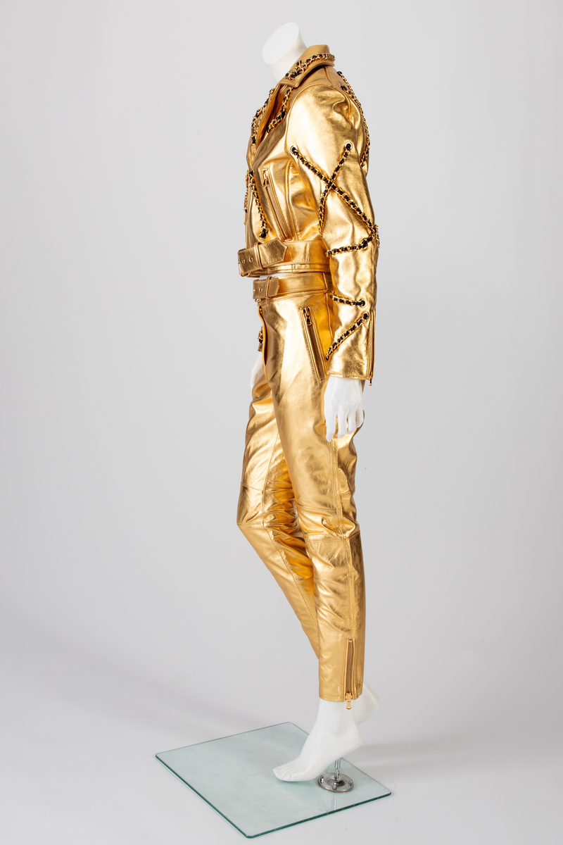 Skinnbukse, jeans-modell, med passepoilerte stikklommer med gullforgylt metallglidelås. På høyre side, nær linningen, en urlomme med klaff og gulltrykknapp. Gylf midt foran med gullforgylt glidelås og trykknapp i linningen. En passepoilert baklomme med gullforgylt glidelås på høyre side. På venstre side er lommen lik urlommen foran. Linningen har fem stikninger. Beltet kommer ut av linningen ved sidesømmen, Beltet har gullforgylt metallspenne og maljer. Formet over kneet. Gullforgylt glidelås med passepoiler ved fotvidden.