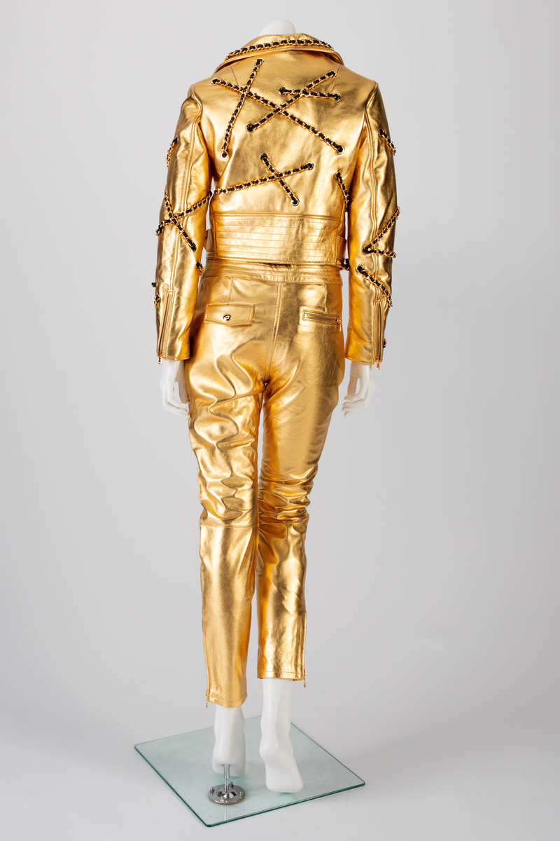 Skinnbukse, jeans-modell, med passepoilerte stikklommer med gullforgylt metallglidelås. På høyre side, nær linningen, en urlomme med klaff og gulltrykknapp. Gylf midt foran med gullforgylt glidelås og trykknapp i linningen. En passepoilert baklomme med gullforgylt glidelås på høyre side. På venstre side er lommen lik urlommen foran. Linningen har fem stikninger. Beltet kommer ut av linningen ved sidesømmen, Beltet har gullforgylt metallspenne og maljer. Formet over kneet. Gullforgylt glidelås med passepoiler ved fotvidden.