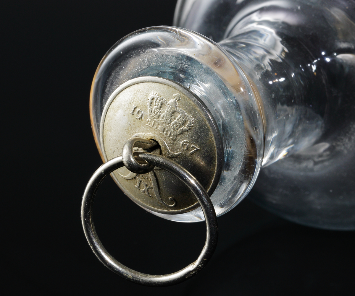 Flaska med två instick som möter varandra i mitten på kroppen. Driven hals med äldre ringförsedd propp med Frederik IX's präglade monogram under en krona och årtalet 1967.