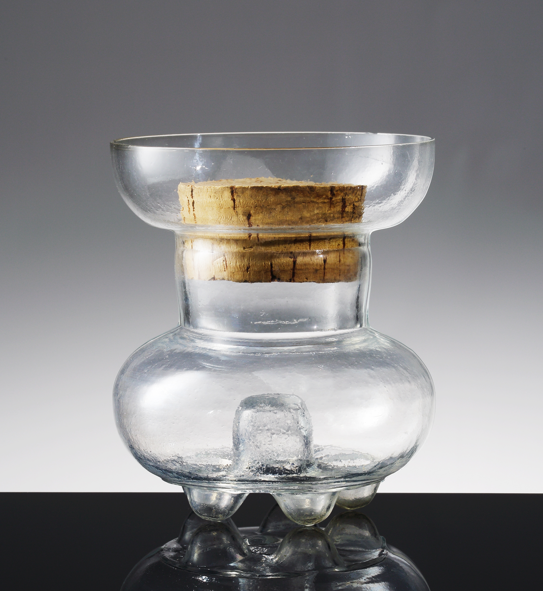 Formblåst timglasformad burk med korkpropp stående på 6 runda fötter med en inskjutande del i mitten.