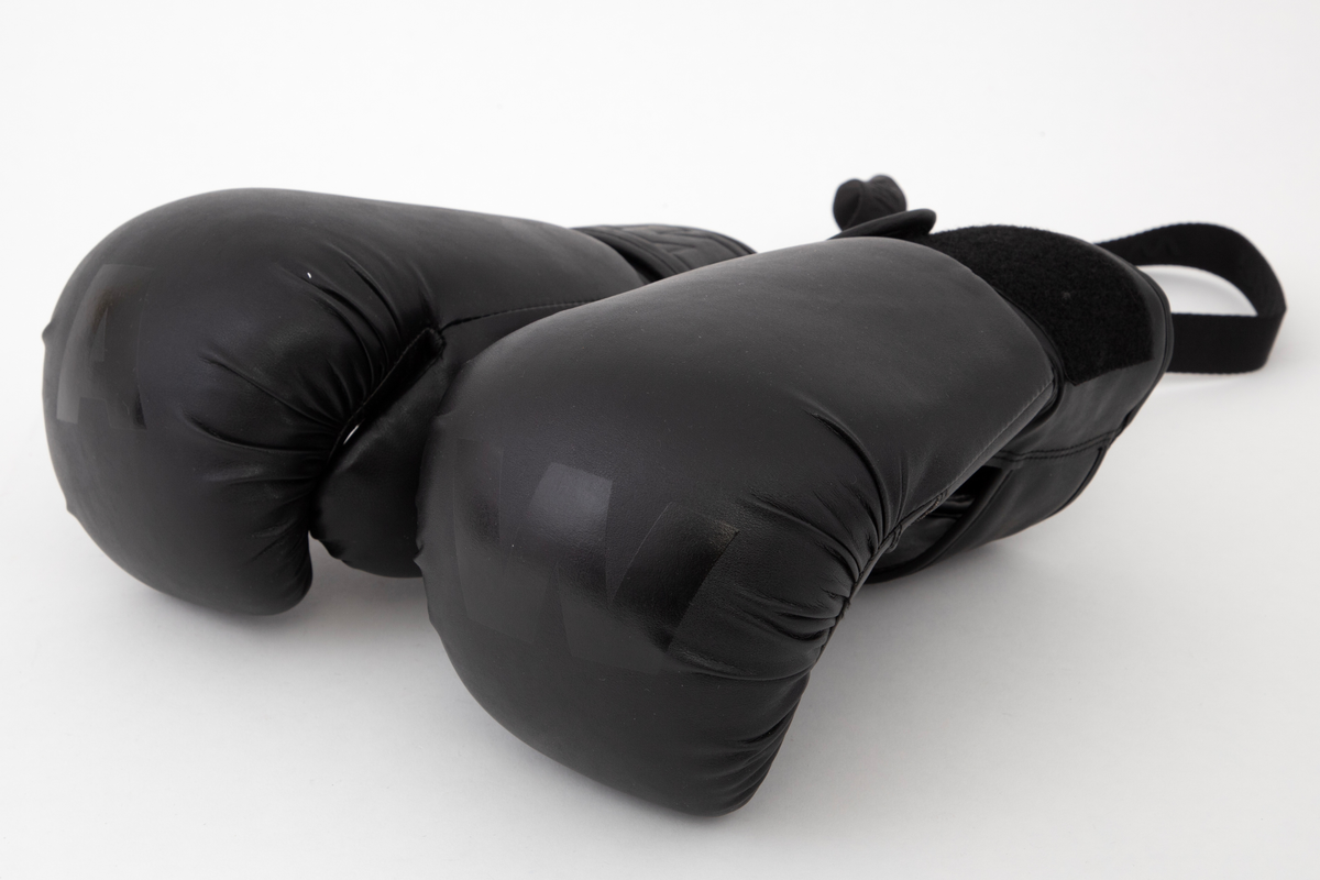 Polstrede og avstivede hansker til bruk ved boksing. Fem små hull i læret sikrer litt ventilasjon inni håndflaten. Lukkes med høy mansjett rundt håndleddet, med åpen splitt på innsiden, ned mot håndflaten. En bred stropp spennes omkring mansjetten og lukkes mot forsiden med like bred borrelås. Denne har bokstavene AW preget inn på forsiden. Ellers har den ene hansken en stor, blank "A" på den knyttede endeflaten, og den andre en tilsvarende "W". Et hull med metallmalje øverst ved splitten på hver hanske brukes til feste for de to krokene i stroppen som binder dem sammen når de ikke er i bruk. Innvendig fôret med syntetisk stoff. Stroppen har navnet WANG vevd inn gjentatte ganger på den ene siden.