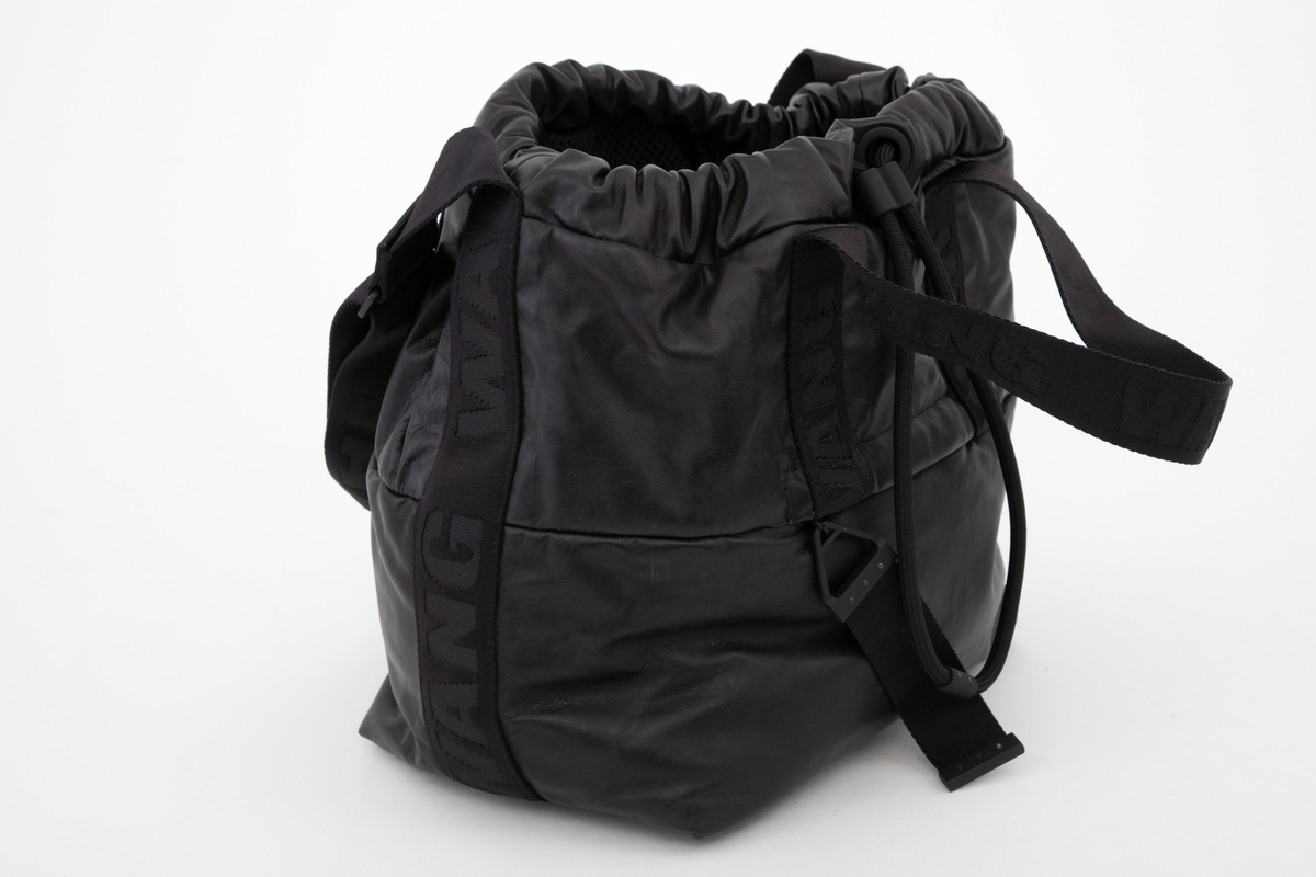 Posete bag av mykt skinn, åpen øverst, men kan lukkes med tykk snor i løpegang. Snoren dras til og stoppes med en "gummihylse". Skulderstroppen er av kraftig, dobbelvevd syntetbånd ivevd WANG. Den kan reguleres ved hjelp av glidespenne i gummiert metall, merket AW, og er fastsydd over sidesømmen helt ned på hver side av vesken. I tillegg har den korte bærehåndtak på for- og bakstykket, festet halveis ned på hver flate. Front- og bakstykket er sammensydd av en øvre og nedre del og skjult i sømmen er det en glidelåslomme på begge sider. Innvendig er det et stort hovedrom. I tillegg er påsydd to mindre lommer på den ene siden og et større glidelåsrom på den andre siden. De utvendige glidelåsene er utstyrt med kraftig drastropp festet i glideren. Denne er av samme bånd ivevd WANG. Vesken er innvendig fôret med nettstoff. Alle lommer og ekstrarom er også sydd av samme nettstoff. Innvendig plate innsydd i veskebunnen, og i tillegg en påsydd gummiplate utvendig, med fire ben av bokstavene AW i høyt relieff.