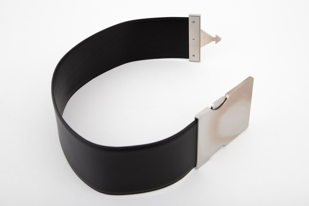 Kraftig, stivt og bredt lærbelte, laminert av to lag og randsydd. Stor beltespenne av børstet stål. Låses ved at en pilformet tapp på det ene, smale beslaget skyves inn i den store "platen", en slags "låskasse", på motsatt side. For å løse ut tappen må to sidetapper på den store platen trykkes inn samtidig. Forsiden på beltespennen er helt glatt, men børstet, mens alle festeskruene kan sees på baksiden. Tilsammen 9 små stjerneskruer fordelt på låsens to endestykker.