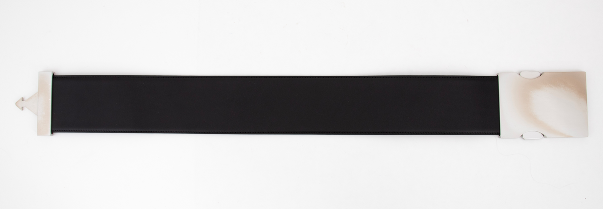 Kraftig, stivt og bredt lærbelte, laminert av to lag og randsydd. Stor beltespenne av børstet stål. Låses ved at en pilformet tapp på det ene, smale beslaget skyves inn i den store "platen", en slags "låskasse", på motsatt side. For å løse ut tappen må to sidetapper på den store platen trykkes inn samtidig. Forsiden på beltespennen er helt glatt, men børstet, mens alle festeskruene kan sees på baksiden. Tilsammen 9 små stjerneskruer fordelt på låsens to endestykker.