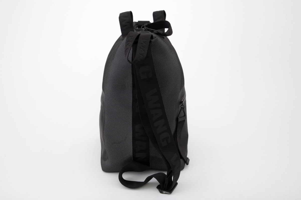 Romslig bag med alternative bæremåter. Enten etter to korte håndtak på øvre kortside, eller etter løkkeformet stropp på enden av øvre kortside, eller etter skulderstropp fra topp til bunn på ene langside. Som skulderbag har sekken også to utforminger: enten bæres den i full størrelse, eller med topp og bunn sammenhektet på motsatt langside. Der er den utstyrt med metallhempe nederst og en slags karabinkrok av metall øverst. Veskens bunn er konstruert med innbrettede kortsider, slik at den ser ut som en konvolutt, og begge langsider og bunnen er forsterket med et svart syntetbånd ivevd gjentatte WANG. Skulderstroppen og alle andre stropper og håndtak er av samme type bånd, de små på toppen en del smalere. Skulderstroppen er dobbel og regulerbar med metallglidestykke merket AW. De to små bærehåndtakene på toppen av bagen er festet på hver side av den gummierte glidelåsen og forsterket med et underliggende bånd av samme type. Bagen er dyp, sydd av et spettet neoprenstoff, svart i bunnfargen og med spetter av sølvfarget, antagelig reflekterende fibre. Alle forsterkningsbånd og stropper er helsvarte. Baggen er fôret med svart nettstoff, og påsydd en stor innerlomme på ene sideveggen. Den er forsterket med smalt bånd av PVC-belagt tekstil og utstyrt med påsydd, matt gummilapp med blank, påtrykt skrift "ALEXANDER WANG X H&M". På for- og baksiden, utvendig, er påsveiset en stor lomme av gummiert tekstil, begge lukket med nesten usynlig, fastsveiset glidelås.