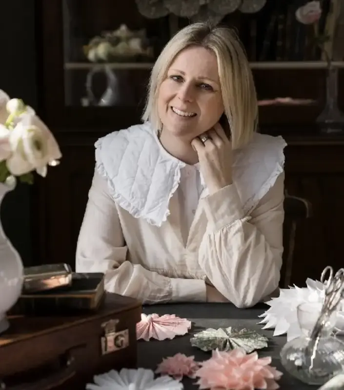 Kvinna i vit tröja sitter vid ett bord med pappersstjärnor i olika färger framför sig.