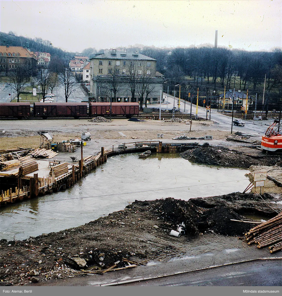 Muddring av Mölndalsån inför planerad byggnation av Mölndals bro. Tid: Omkring 1974. I bakgrunden syns Kvarnbygatan 1 (i folkmun kallad Röda kvarn eller Moulin Rouge) och Kvarnbyskolan syns till vänster i bild. Bakom Röda kvarn sticker den ljusa byggnad som har inrymt ett apotek och haft adressen Kvarnbygatan 9. Dessutom syns den skorsten som funnits länge på pappersbruket Papyrus område, i bakgrunden, till höger räknat.
Några godstågvagnar syns på järnvägsspåret.