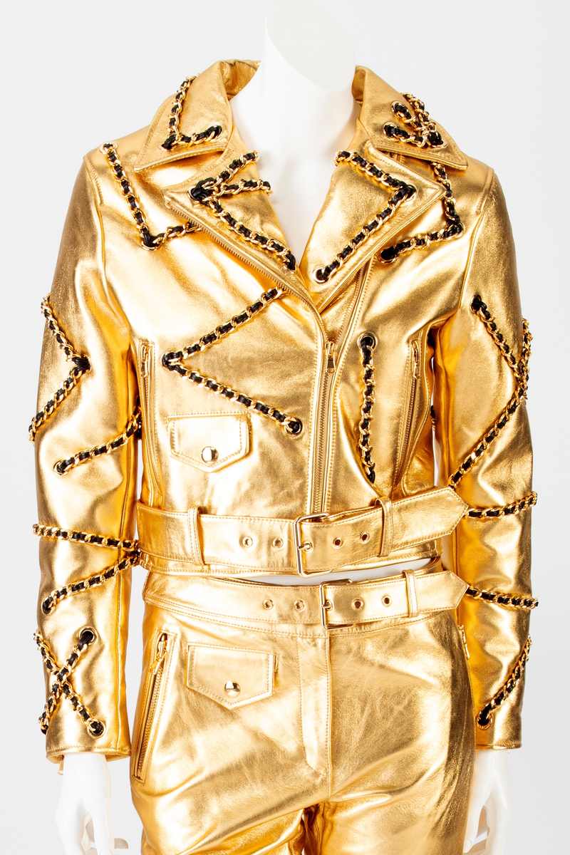 Kort skinnjakke med passepoilerte lommer med gullfarget glidelås og liten lomme med klaff, krage og slag. Lukkes med glidelås midt foran. Tosøms erme med smalt glidelås ved håndleddet. Jakken er dekorert med gullfarget kjetting med skinnsnor smettet inn, tredd gjennom gullfargede maljer på plagget. Dekoren er på krage, forstykke, ermer og bakstykke. Belte med gullfarget spenne i jakkens nederkant.