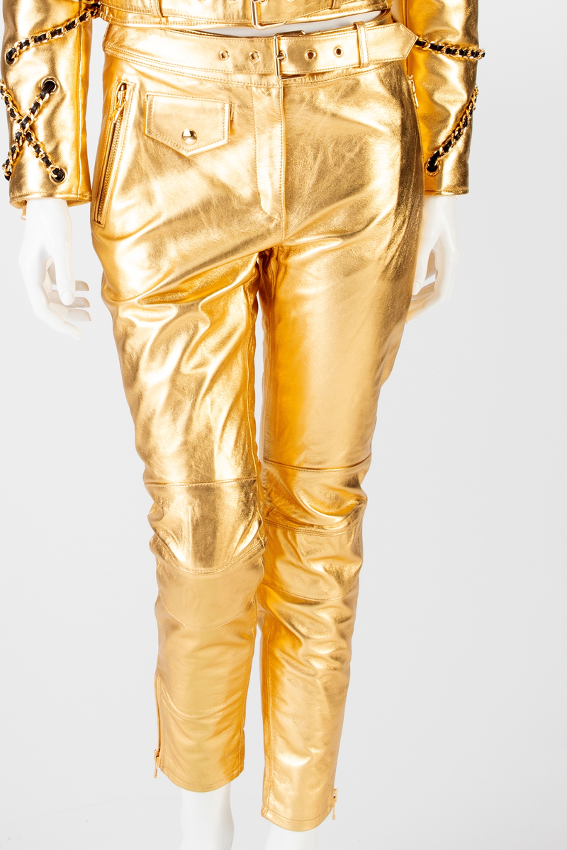 Skinnbukse, jeans-modell, med passepoilerte stikklommer med gullforgylt metallglidelås. På høyre side, nær linningen, en urlomme med klaff og gulltrykknapp. Gylf midt foran med gullforgylt glidelås og trykknapp i linningen. En passepoilert baklomme med gullforgylt glidelås på høyre side. På venstre side er lommen lik urlommen foran. Linningen har fem stikninger. Beltet kommer ut av linningen ved sidesømmen, Beltet har gullforgylt metallspenne og maljer. Formet over kneet. Gullforgylt glidelås med passepoiler ved fotvidden.