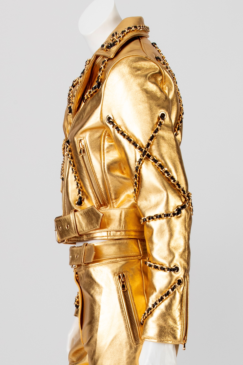 Kort skinnjakke med passepoilerte lommer med gullfarget glidelås og liten lomme med klaff, krage og slag. Lukkes med glidelås midt foran. Tosøms erme med smalt glidelås ved håndleddet. Jakken er dekorert med gullfarget kjetting med skinnsnor smettet inn, tredd gjennom gullfargede maljer på plagget. Dekoren er på krage, forstykke, ermer og bakstykke. Belte med gullfarget spenne i jakkens nederkant.